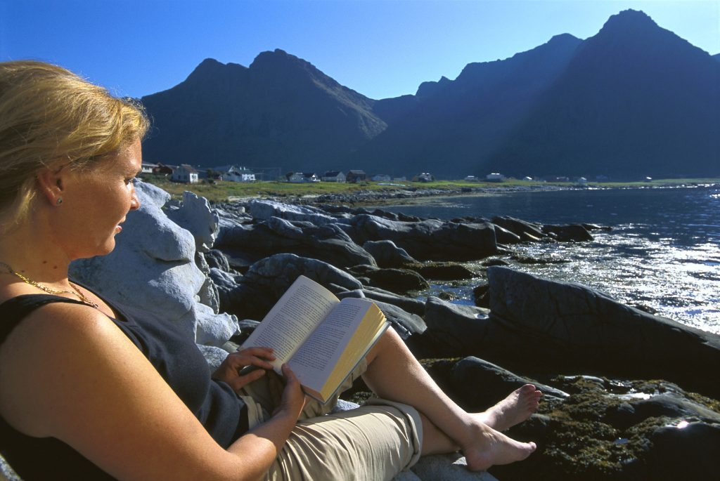 Kvinne leser bok i fjæra utenfor et tettsted i Nordland.